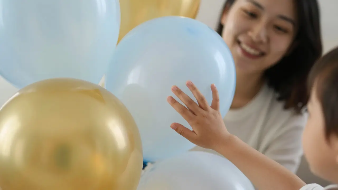 A child's hand gently touching a cluster of translucent balloons, with a smiling adult in the blurred background.